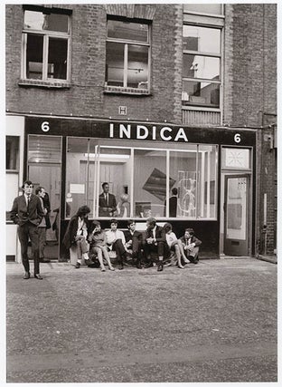 Item #41263 A b/w photograph by Keith Avison of a group of people sitting against the front wall...
